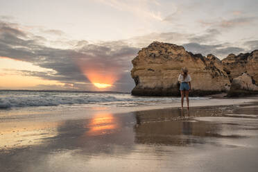 Rückenansicht einer nicht erkennbaren Frau in Freizeitkleidung und Hut, die bei Sonnenuntergang in den Benagil-Höhlen an der Algarve, Portugal, am Meer steht - ADSF20872