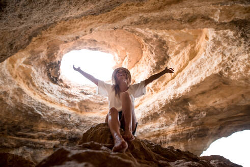 Von unten: fröhliche Frau mit Hut, die mit ausgestreckten Armen auf einem Felsen sitzt und auf ein Loch in der Decke einer Höhle in der Nähe des Meeres schaut, an einem sonnigen Tag an der Algarve, Portugal - ADSF20873