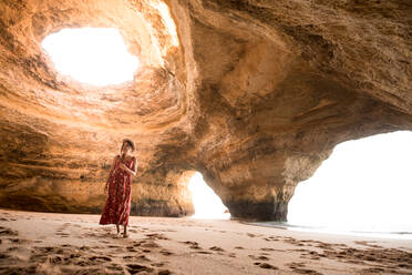 Frau läuft auf Sand in einer geräumigen Steinhöhle in Richtung Meer an einem sonnigen Tag in den Benagil-Höhlen an der Algarve, Portugal - ADSF20874