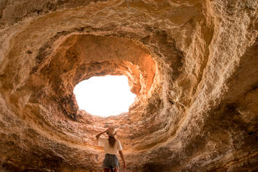 Rückenansicht einer anonymen Frau mit Hut, die ein Loch in der Decke einer Höhle in der Nähe des Meeres an einem sonnigen Tag an der Algarve, Portugal, betrachtet - ADSF20875