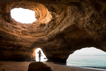 Rückenansicht einer anonymen Frau mit Hut, die in einem Felsen steht und auf ein Loch in der Decke einer Höhle in der Nähe des Meeres blickt, an einem sonnigen Tag an der Algarve, Portugal - ADSF20876