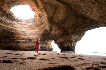 Rückenansicht einer nicht erkennbaren Frau, die in einer geräumigen Steinhöhle auf Sand in Richtung Meer läuft, an einem sonnigen Tag in den Benagil-Höhlen an der Algarve, Portugal - ADSF20877