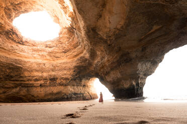 Back view of unrecognizable female walking on sand inside spacious stone cave towards sea on sunny day in Benagil Caves in Algarve, Portugal - ADSF20878