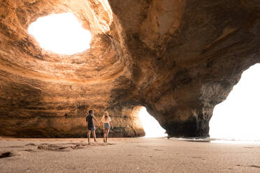 Rückansicht eines glücklichen Mannes und einer glücklichen Frau, die sich an den Händen halten, während sie in der Höhle von Benagil in der Algarve, Portugal, über den Sand laufen - ADSF20879