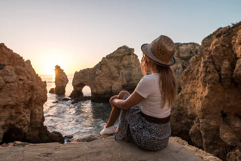 Seitenansicht einer anonymen Frau mit Hut, die auf einem Stein sitzt und das Meer und die Klippen bei Sonnenuntergang in Ponta da Piedade an der Algarve, Portugal, bewundert - ADSF20882