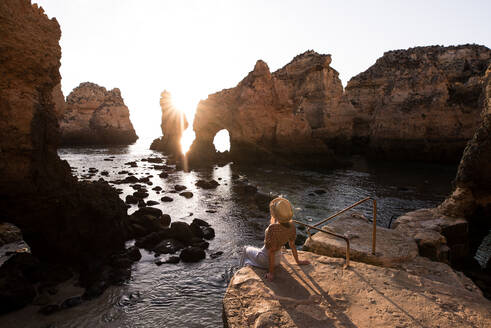 Anonyme Frau mit Hut sitzt auf einem Stein und bewundert das Meer und die Klippen bei Sonnenuntergang in Ponta da Piedade an der Algarve, Portugal - ADSF20884