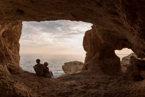 Baxk-Ansicht eines Mannes und einer Frau, die im Eingang einer Felsenhöhle in der Nähe des Meeres in den Algar seco-Höhlen an der Algarve, Portugal, sitzen - ADSF20889