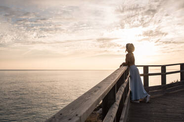 Side view of unrecognizable female traveler in summer outfit and straw hat relaxing on wooden pier and admiring waving sea against cloudy sunset sky - ADSF20890