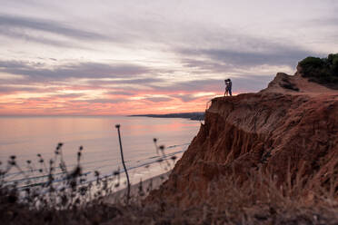 Seitenansicht eines unerkennbaren Paares, das zusammen in einer Bergklippe an der Küste steht, während eines schönen Sonnenuntergangs in Tavira an der Algarve, Portugal - ADSF20895