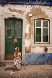 Junge, fröhliche Reisende in legerer Sommerkleidung, die auf der Straße vor der Tür eines alten Gebäudes in Algarve, Portugal, sitzt und wegschaut - ADSF20899