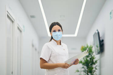 Eine Dermatologin in Uniform und medizinischer Maske steht mit einem Klemmbrett im Flur eines modernen Schönheitszentrums und schaut in die Kamera - ADSF20907
