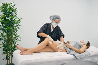 Masseurin in Maske und Handschuhen bei der Massage der Beine einer Kundin, die auf einem Tisch liegt, während der Körperpflege in einem Schönheitszentrum - ADSF20915