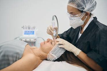 Dermatologist wearing protective face mask using machine for cleansing pores on face of mature female lying on table in beauty center during covid 19 pandemic - ADSF20916