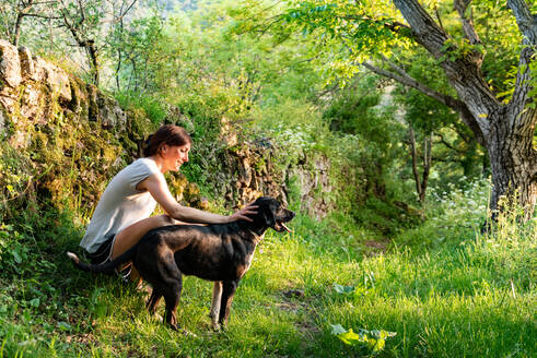 Seitenansicht der zufriedenen Besitzerin, die ihren liebenswerten treuen Hund streichelt, während sie sich gemeinsam an einem sonnigen Tag im grünen Wald entspannt - ADSF20917