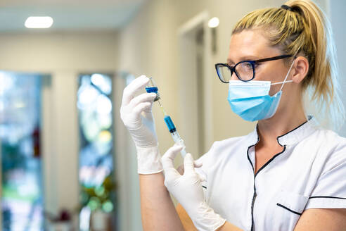 Ärztin mit Maske und Latexhandschuhen füllt Spritze mit flüssigem Medikament bei der Arbeit im Krankenhaus - ADSF20919