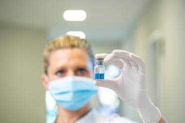 Sanitäterin mit Schutzmaske und Handschuhen steht mit einer blauen chemischen Flüssigkeit in einem Glasfläschchen in einem Krankenhaus während einer Pandemie des Coronavirus - ADSF20920