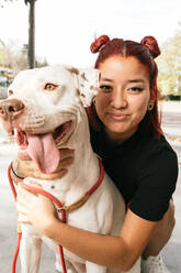 Lächelnde junge asiatische Besitzerin, die einen gehorsamen reinrassigen American Pit Bull Terrier Hund umarmt und in die Kamera schaut - ADSF20932