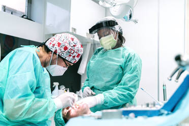 African American nurse in protective uniform and face shield assisting professional dentist in face mask during procedure with teeth of patient in modern stomatology clinic during coronavirus pandemic - ADSF20937
