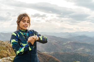 Selbstbewusste junge ethnische Sportlerin in Sportkleidung, die beim Training im Freien in bergigem Gelände gegen den bewölkten Himmel die Zeit auf ihrer Smartwatch überprüft - ADSF20938