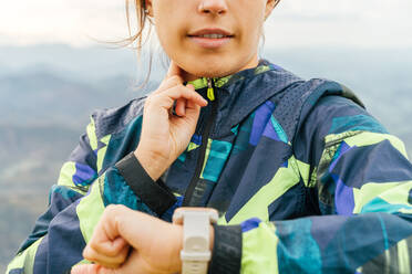 Selbstbewusste junge ethnische Sportlerin in Sportkleidung, die beim Training im Freien in bergigem Gelände gegen den bewölkten Himmel die Zeit auf ihrer Smartwatch überprüft - ADSF20939