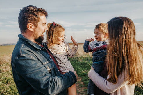 Seitenansicht der Mutter und des Vaters, die auf einem Feld stehen, mit süßen kleinen Geschwistern, die sich gegenseitig High Five geben, während eines Wochenendes auf dem Lande - ADSF20958