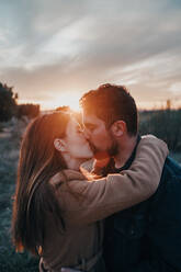Zartes Paar, das sich mit geschlossenen Augen küsst, während es auf einem Feld mit Kind steht und das Wochenende vor dem Hintergrund des Sonnenuntergangs genießt - ADSF20971