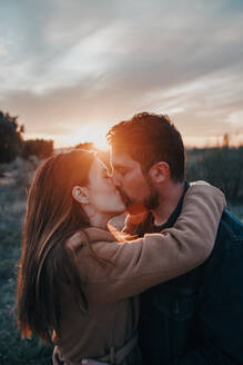 Tender couple kissing with closed eyes while standing in field with child and enjoying weekend on background of sunset sky - ADSF20971