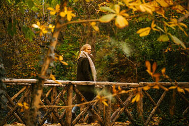 Side view of tranquil female in outerwear walking along wooden bridge and admiring scenery of woods in fall - ADSF20975