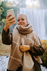 Fröhliche Frau in Oberbekleidung, die in einem herbstlichen Wald steht und ein Selfie mit ihrem Smartphone auf dem Hintergrund eines beeindruckenden Wasserfalls macht - ADSF20977