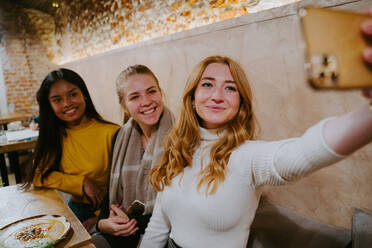 Group of cheerful diverse female friends sitting at table in cafe and taking selfie on smartphone while spending weekend together - ADSF20984