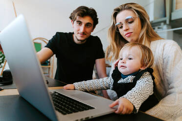 Young couple and toddler sitting at table and watching movie together while spending weekend at home - ADSF20986