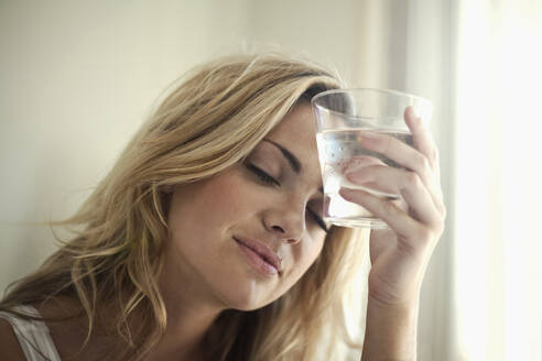 Young woman holding glass of water against her forehead at home - AJOF01041