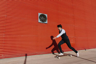 Geschäftsmann mit Longboard, der auf dem Gehweg an der Mauer Skateboard fährt - EGAF01753