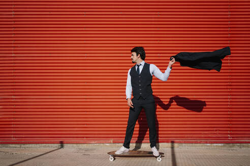 Male entrepreneur holding blazer jacket while standing on longboard against wall - EGAF01754