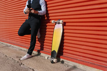 Geschäftsmann, der ein Mobiltelefon auf einem Longboard benutzt und sich an eine orangefarbene Wand lehnt - EGAF01757