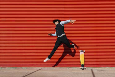 Male entrepreneur jumping by longboard against wall - EGAF01758