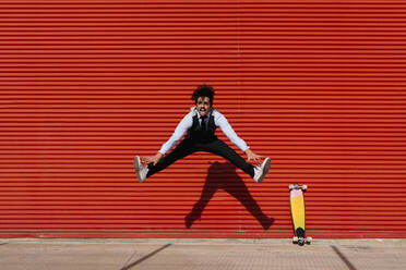 Playful businessman jumping by skateboard against wall - EGAF01759