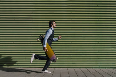 Männlicher Unternehmer mit Longboard läuft auf dem Fußweg an der grünen Wand - EGAF01763