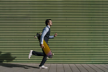 Male entrepreneur with longboard running on footpath by green wall - EGAF01763