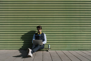 Geschäftsmann, der einen Laptop benutzt und auf einem Skateboard vor einer grünen Wand sitzt - EGAF01764