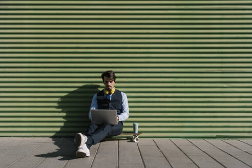 Businessman using laptop while sitting on skateboard against green wall - EGAF01764