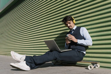 Businessman with laptop holding coffee cup while sitting on longboard against green wall - EGAF01765