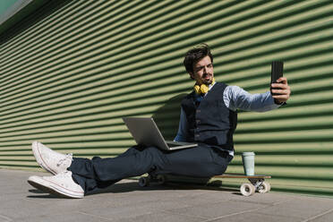 Junger Geschäftsmann mit Laptop, der ein Selfie macht, während er auf einem Skateboard an der Wand sitzt - EGAF01766