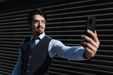 Lächelnder Geschäftsmann, der ein Selfie macht, während er an einer schwarzen Wand steht - EGAF01774