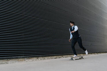 Lächelnder Geschäftsmann, der an einer schwarzen Wand Skateboard fährt - EGAF01779