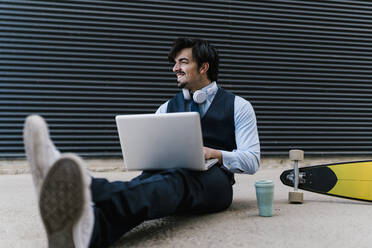 Smiling handsome businessman with laptop looking away while sitting on footpath against wall - EGAF01783