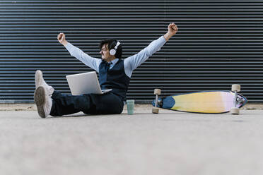 Carefree businessman with laptop sitting on footpath against wall - EGAF01784