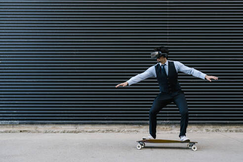 Geschäftsmann, der auf einem Longboard Skateboard fährt, während er einen Film in einem Virtual-Reality-Simulator an einer schwarzen Wand betrachtet - EGAF01785