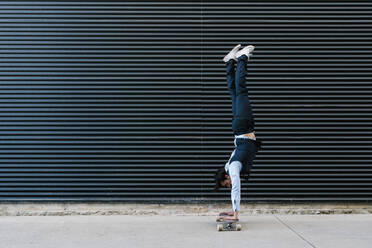 Männlicher Unternehmer macht Handstand auf Longboard vor schwarzer Wand - EGAF01787