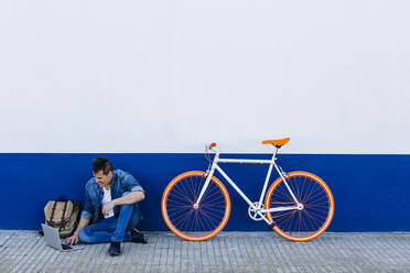 Smiling man using laptop by bicycle while sitting on footpath against wall - XLGF01184